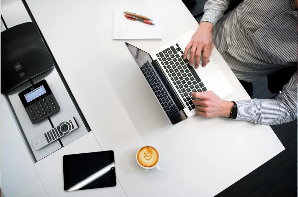 A desk seen from above with a laptop, a notebook, a phone and a coffee.